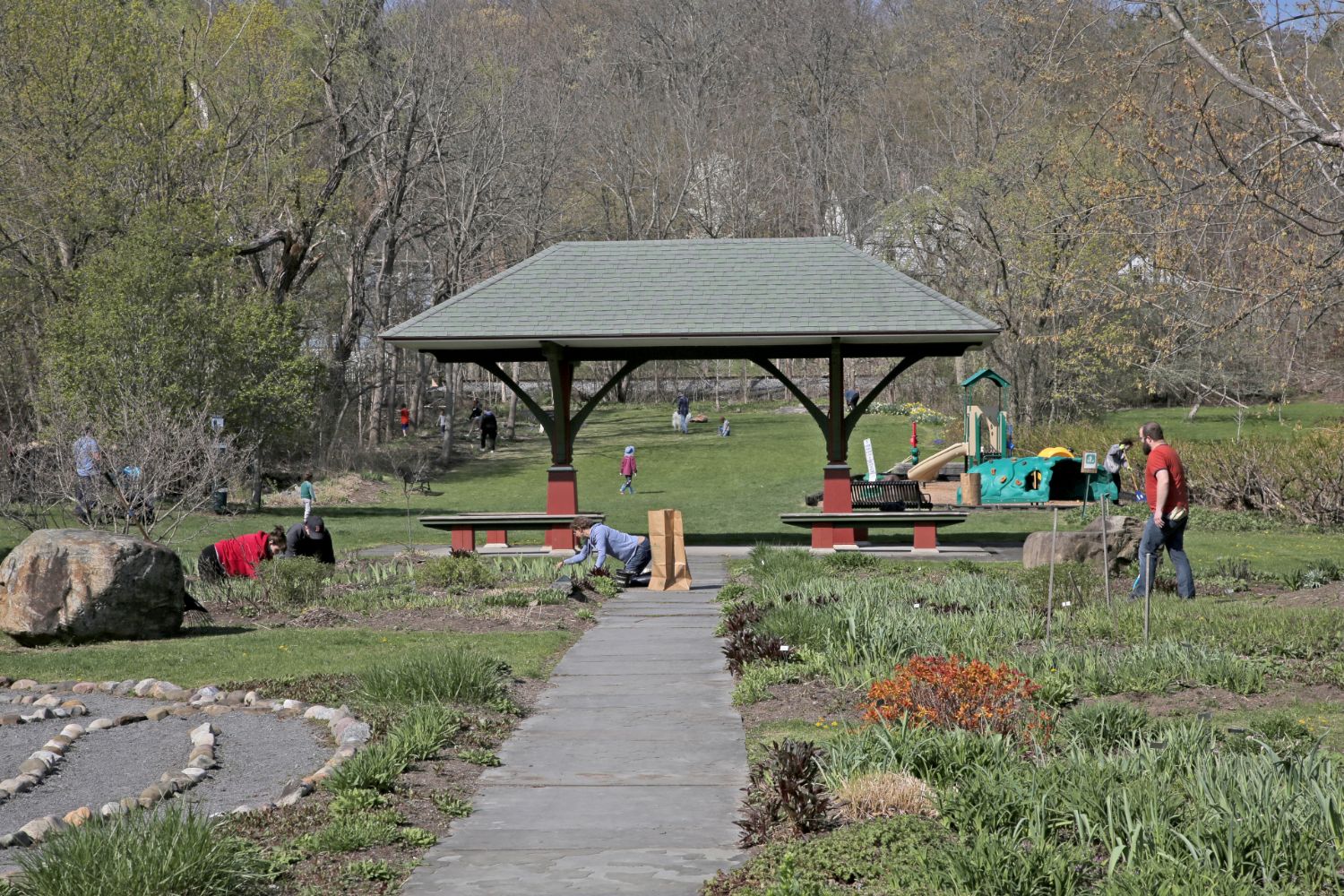 Volunteers tend garden beds near a park pavilion and playground in spring