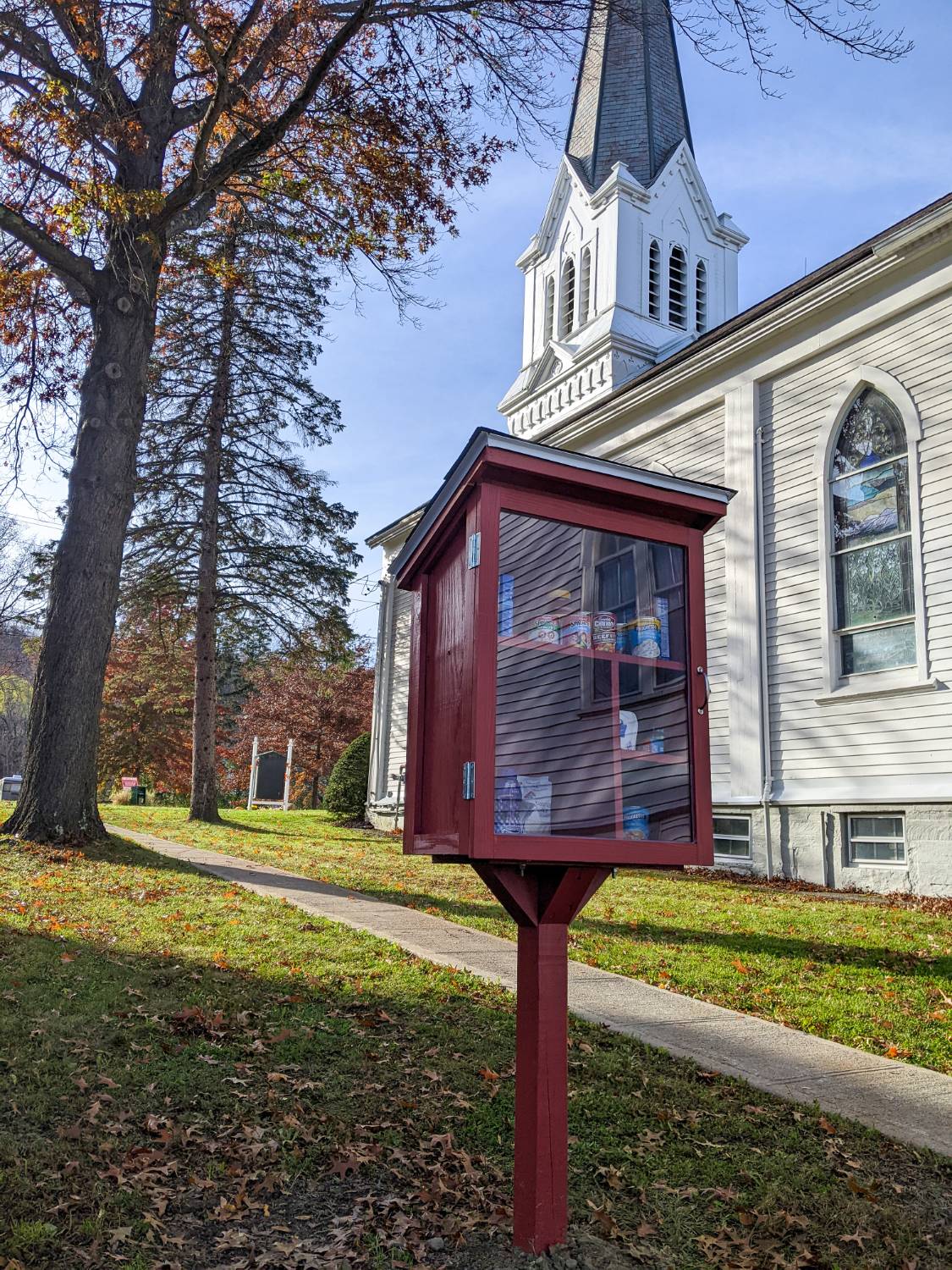 A red community food pantry box beside a white church on an autumn day